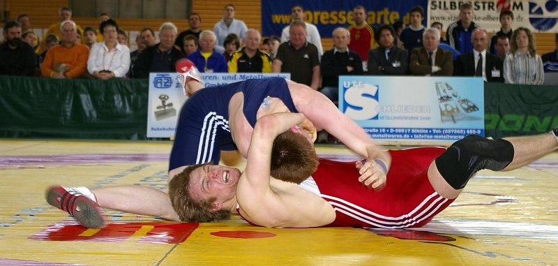 DM_OlbernhauSonntagFinalkaempfe021.jpg - Im Finale der zweithöchsten Gewichtsklasse 85 kg hätte man beinahe die (in hohen Altersklassen) seltenste Art und Weise gesehen, einen Kampf zu gewinnen.Michael Rips (rot, Nordrhein-Westfalen) versuchte hier zwar unter vollster Anstrengung eine Technik, aber sein Genger konterte und hätte ihn beinahe auf die Schultern gelegt. Sind nämlich beide Schultern für eine Sekunde auf der Matte, so ist der Kampf (und nicht nur die Runde) verloren.In diesem Falle geschah dies zwar nicht, der Sieg ging nach drei Runden aber dennoch an Sebastian Wendel (blau, Thüringen).